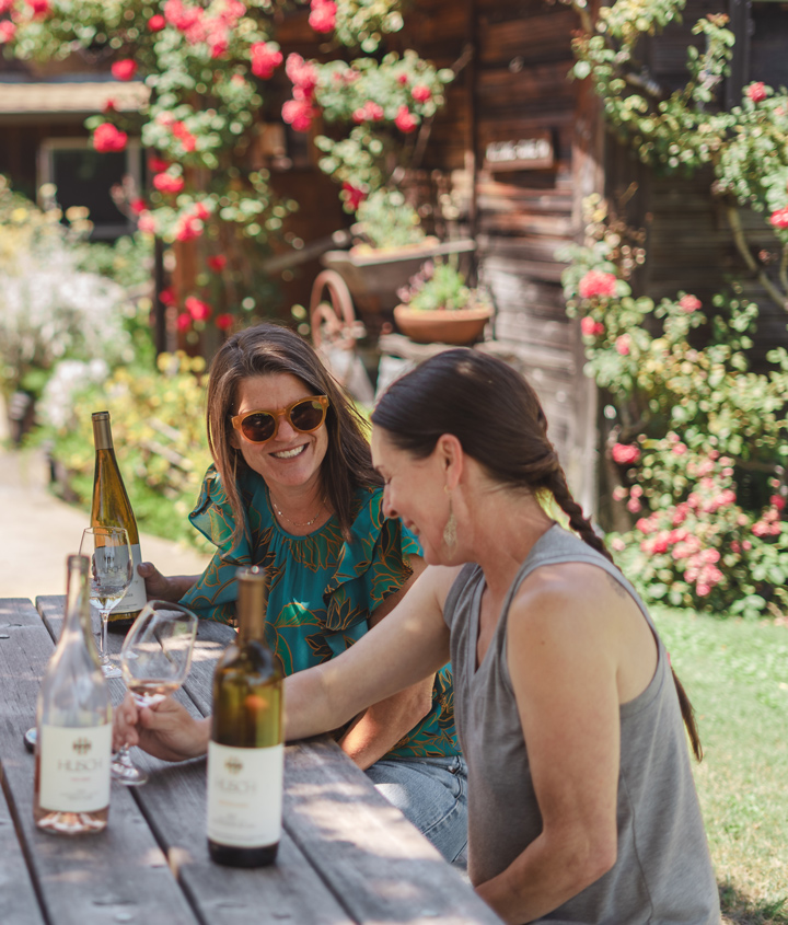 Two women share a laugh while sitting at a picnic table, enjoying a glass of wine.