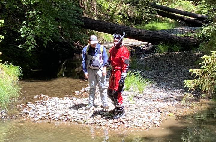 Two scientists, one in a wetsuit with a snorkel, survey the river from the bank.